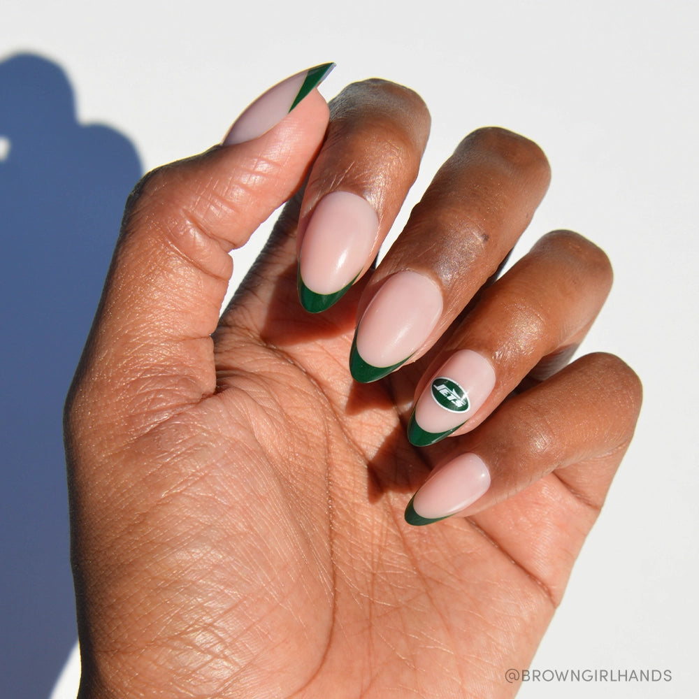 A hand with medium brown skin wears glamnetic’s New York Jets Press-On Nail Set: almond nails with a nude base, green French tips, and a green and white striped oval design on the ring finger. The background is white.