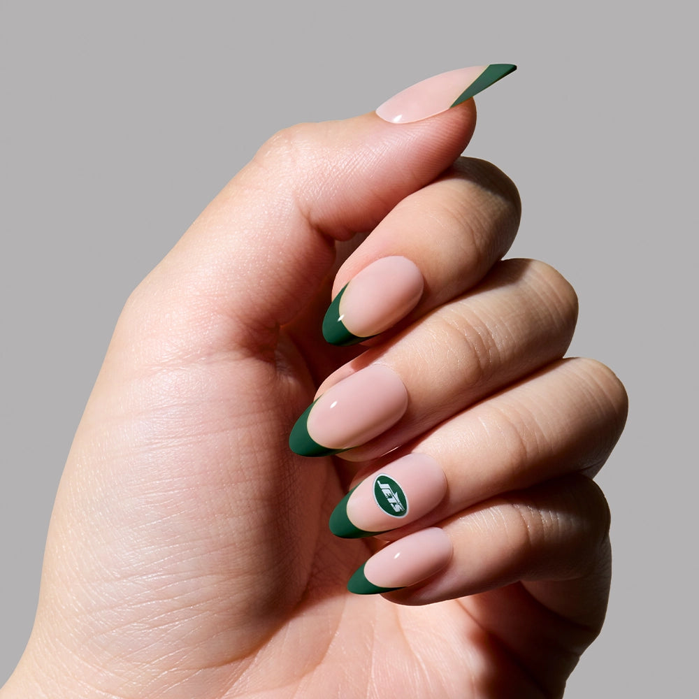 A hand with almond French tips flaunts nude polish and dark green tips from the glamnetic New York Jets Press-On Nail Set; one nail features a white leaf design on the green tip, set against a neutral gray background. - Product image