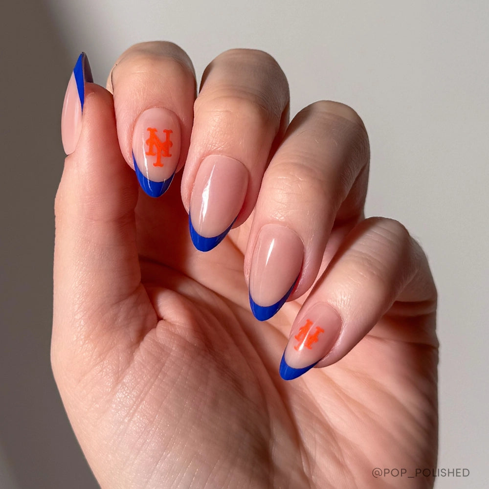 A hand displays the glamnetic New York Mets Press-On Nail Set with almond-shaped nails, blue French tips, and orange NY logos on two nails—all inspired by the Mets. The plain, well-lit background highlights this trendy nail design.