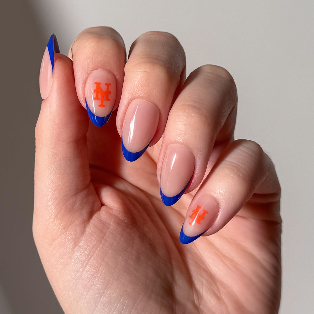 A hand showcases almond-shaped nude nails featuring blue French tips and orange New York Mets logos on the middle and pinky fingers, styled with glamnetic’s MLB Press-On Nails set against a plain light gray background. - Product image