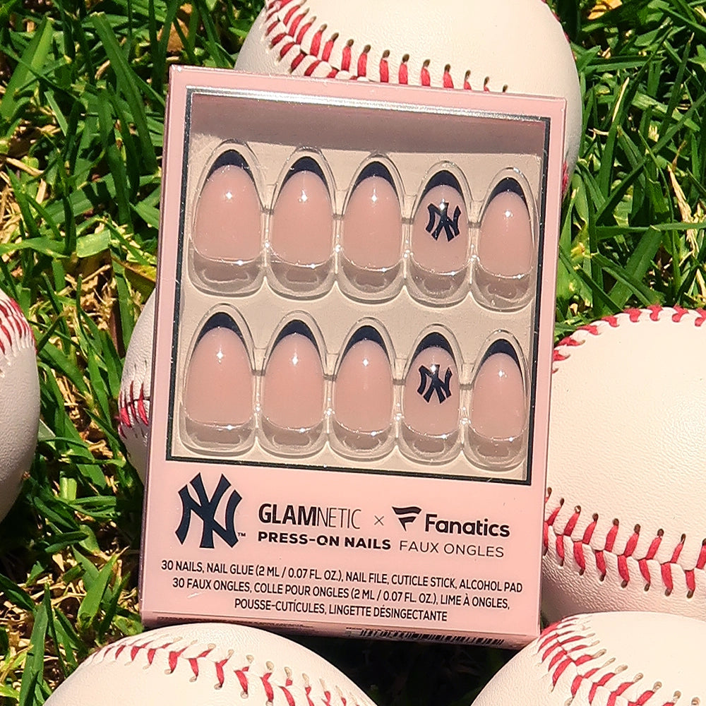 The glamnetic New York Yankees Press-On Nail Set features salon-quality nails with a nude base, blue tips, and a Yankees logo accent nail, perfect for fans. Displayed on grass and surrounded by baseballs. - Product image