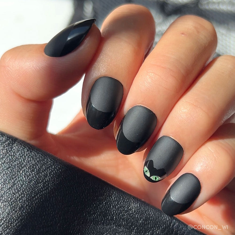 A hand with matte black almond-shaped nails, featuring glossy tips and a nail with a cat face and green eyes—perfect for fans of glamnetic’s Nine Lives set or anyone who loves chic black cat nails.