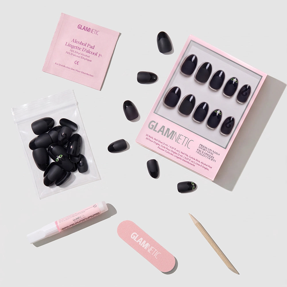 A set of Nine Lives short oval press-on nails in black by glamnetic, along with glue, a wooden cuticle stick, nail file, alcohol pad, and extra nails, are arranged neatly on a light gray surface. - Product image