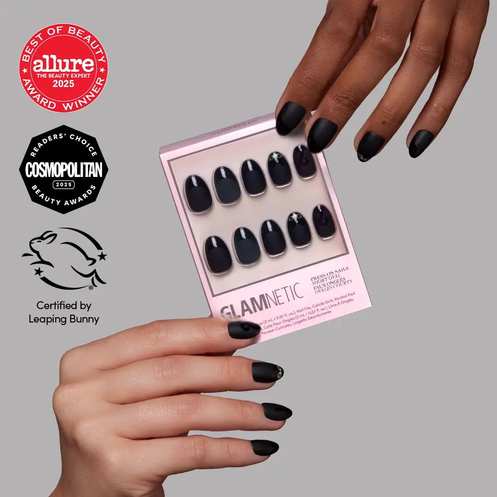 Two hands with black matte glamnetic press-on nails in the “Nine Lives” style hold a pink glamnetic nail set box showcasing salon-quality nails. Beauty award logos and a cruelty-free certification are visible on the left side of the image.