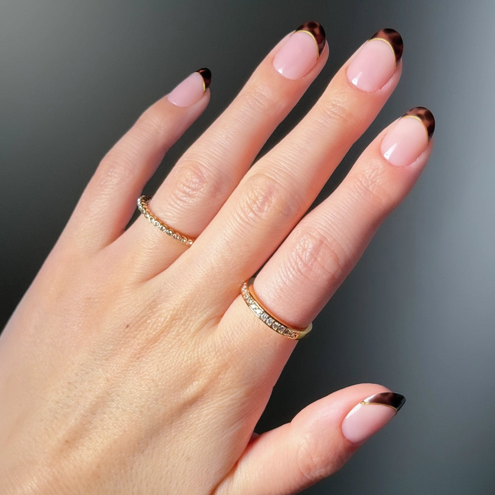 A hand with light skin tone wears glamnetic’s No Apologies gold rings and chic press-on nails, featuring short round manicured nude nails with dark brown tortoiseshell French tips, set against a softly blurred gray background.