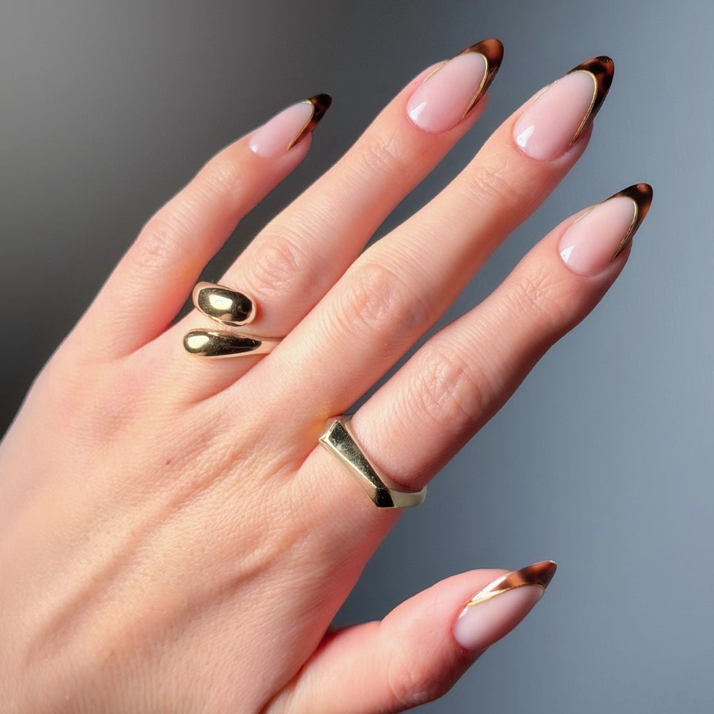 A hand with almond-shaped nails in glamnetic’s No Competition—a nude base with tortoiseshell French tips—adorned with two gold rings (a simple band and an open, curved design) against a blurred background. - Product image