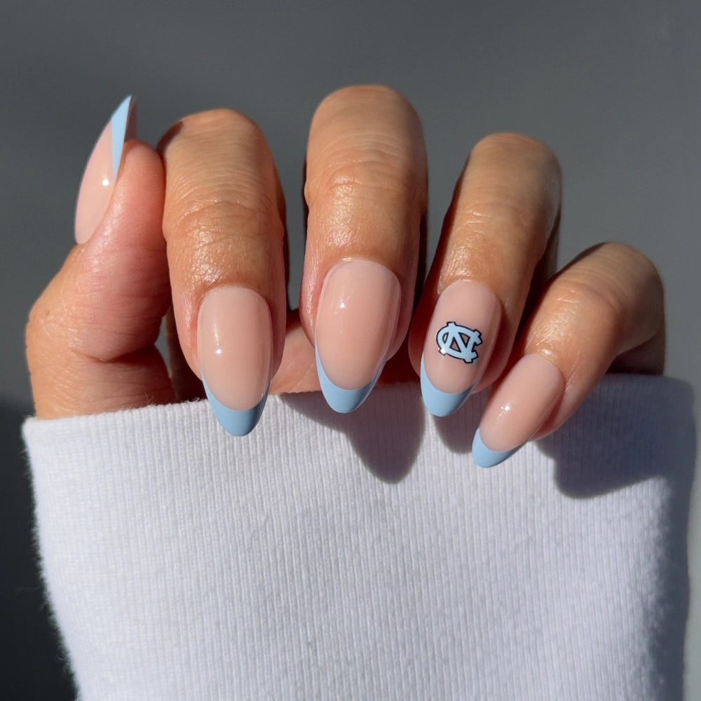 A hand models glamnetic’s North Carolina Tar Heels Press-On Nail Set: almond-shaped, salon-quality nails with pale nude polish, Carolina-blue French tips, and a UNC logo on the ring finger. The model wears a white long-sleeve top.