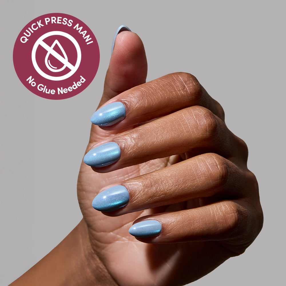 A hand with neatly manicured, metallic blue glamnetic Ocean Prism press-on nails is shown against a gray background. A circular label reads Quick Press Mani, No Glue Needed, with a crossed-out drop icon to highlight their ease of use. - Product image