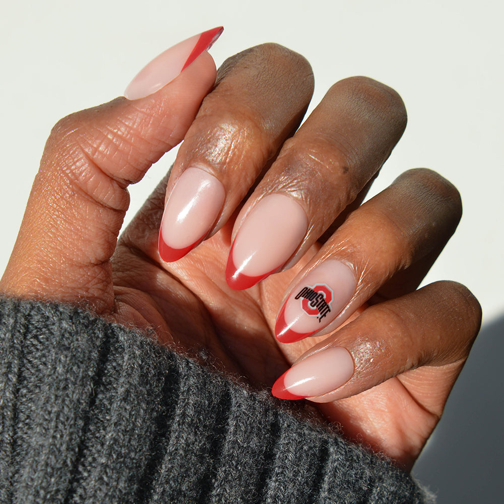 A hand wearing glamnetic Collegiate Press-On Nails showcases almond-shaped tips with a nude base, bold red French accents, and an Ohio State logo on the ring finger. The person is dressed in a gray ribbed sweater. - Product image