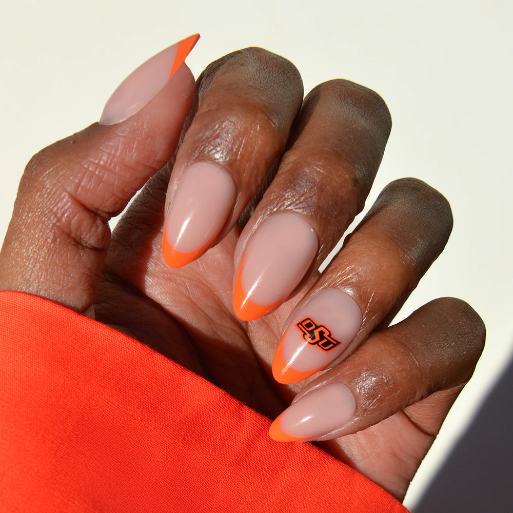 A hand showcases glamnetic Collegiate Press-On Nails with almond-shaped tips, featuring a nude base, vibrant orange French tips, and an OSU logo on the ring finger. The person is wearing an orange sleeve. - Product image