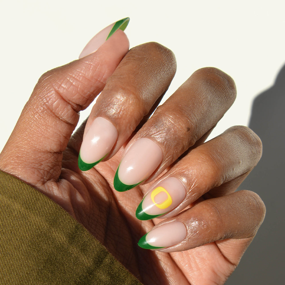 A hand with almond-shaped nails showcases glamnetic’s Collegiate Press-On Nails, featuring a nude base, green French tips, and a yellow square outline accent on the ring finger. The person is wearing an olive green sleeve. - Product image