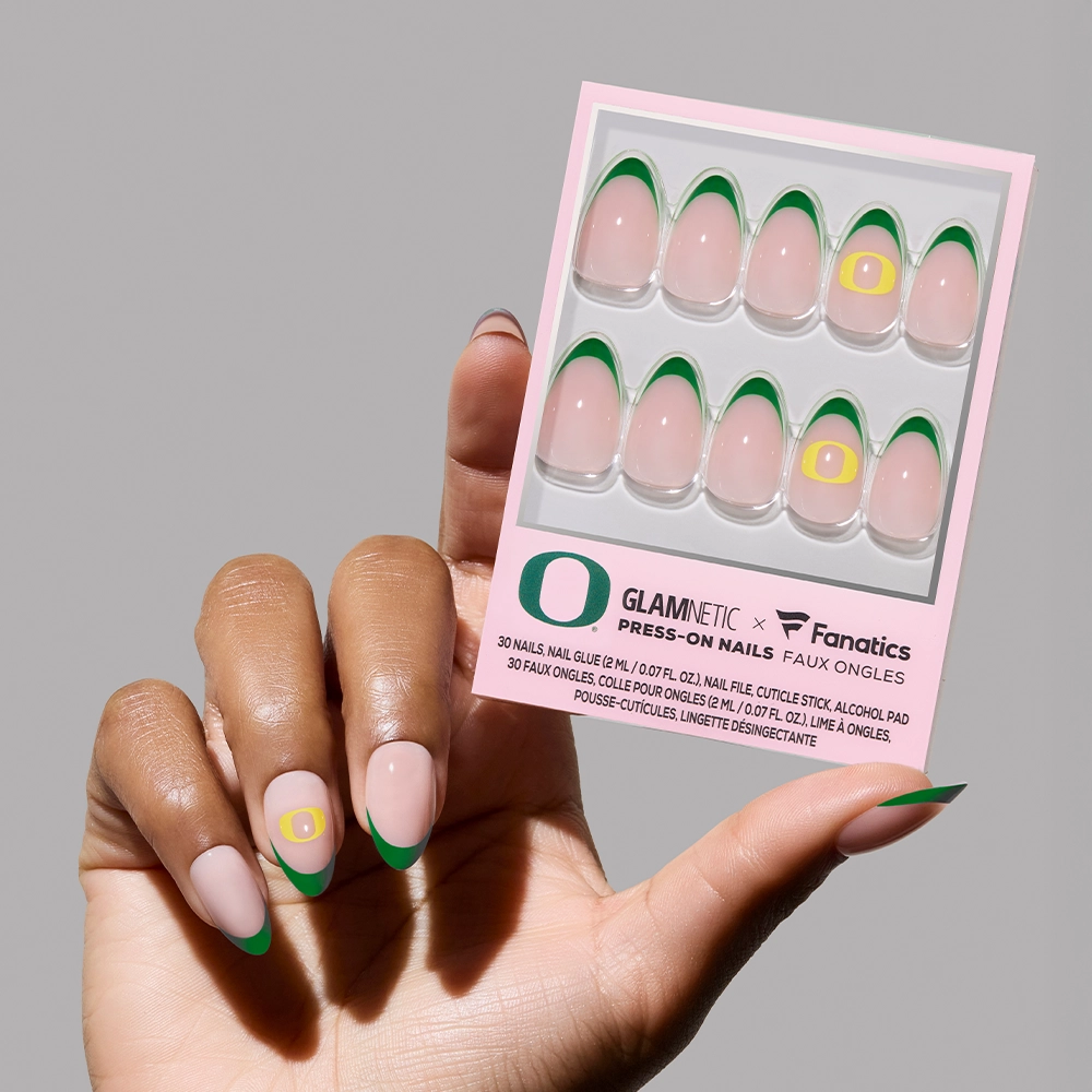 A hand with manicured green French tip nails, featuring the Oregon Ducks logo on the ring finger, holds a box of glamnetic Oregon Ducks Press-On Nail Set—a vibrant collegiate team logo nail set. - Product image
