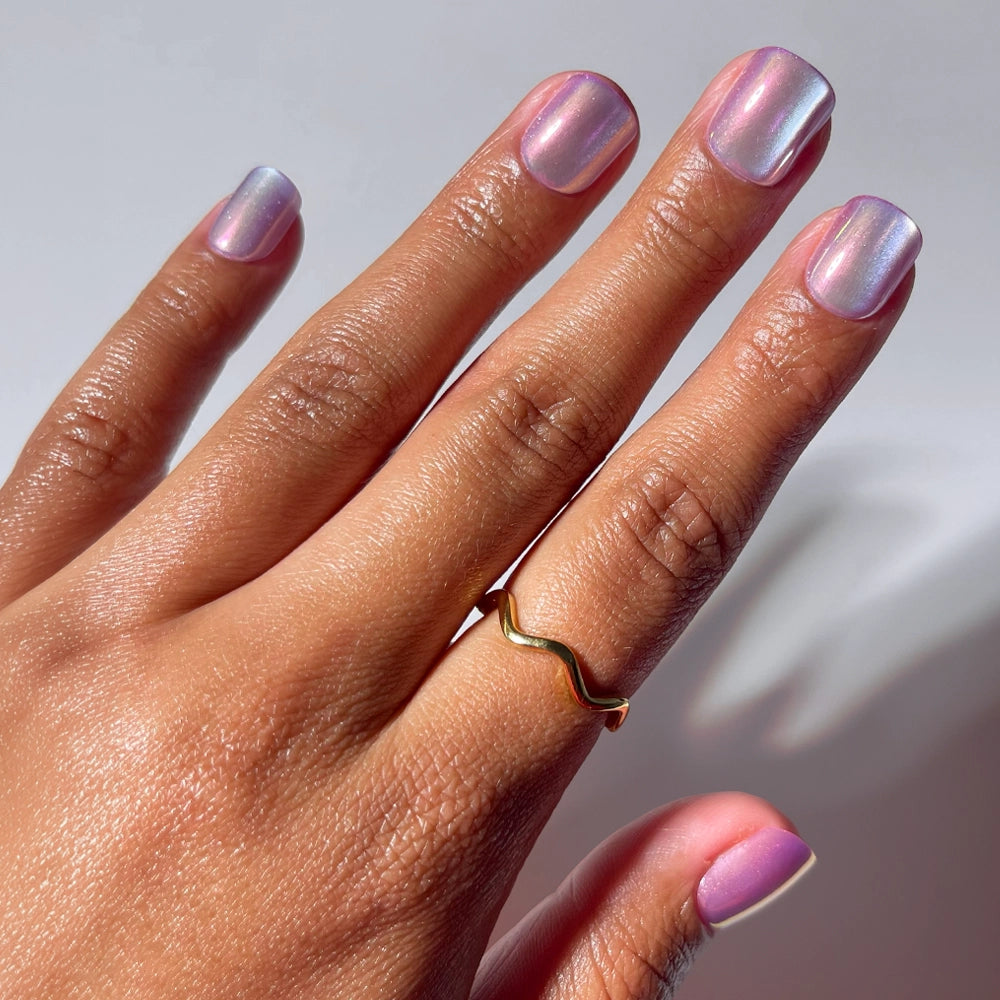 A hand with light brown skin wears a gold wavy ring and glamnetic's Oyster Baby pearlescent press-on nails in shiny, iridescent lilac on the middle finger. The softly blurred, light background highlights the look.