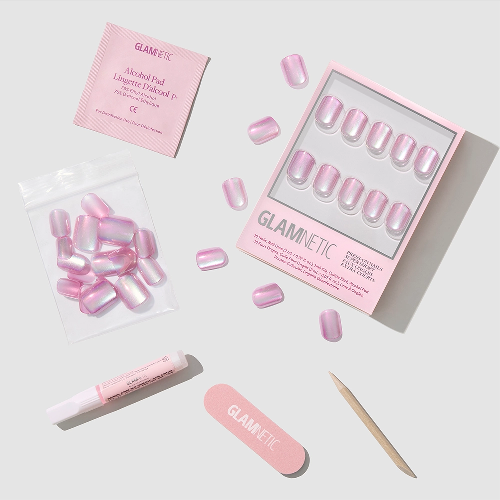 Oyster Baby Press-On Nails by glamnetic in a pink iridescent finish come with an alcohol pad, nail file, cuticle stick, adhesive tube, and storage bag—all displayed on a light gray surface.