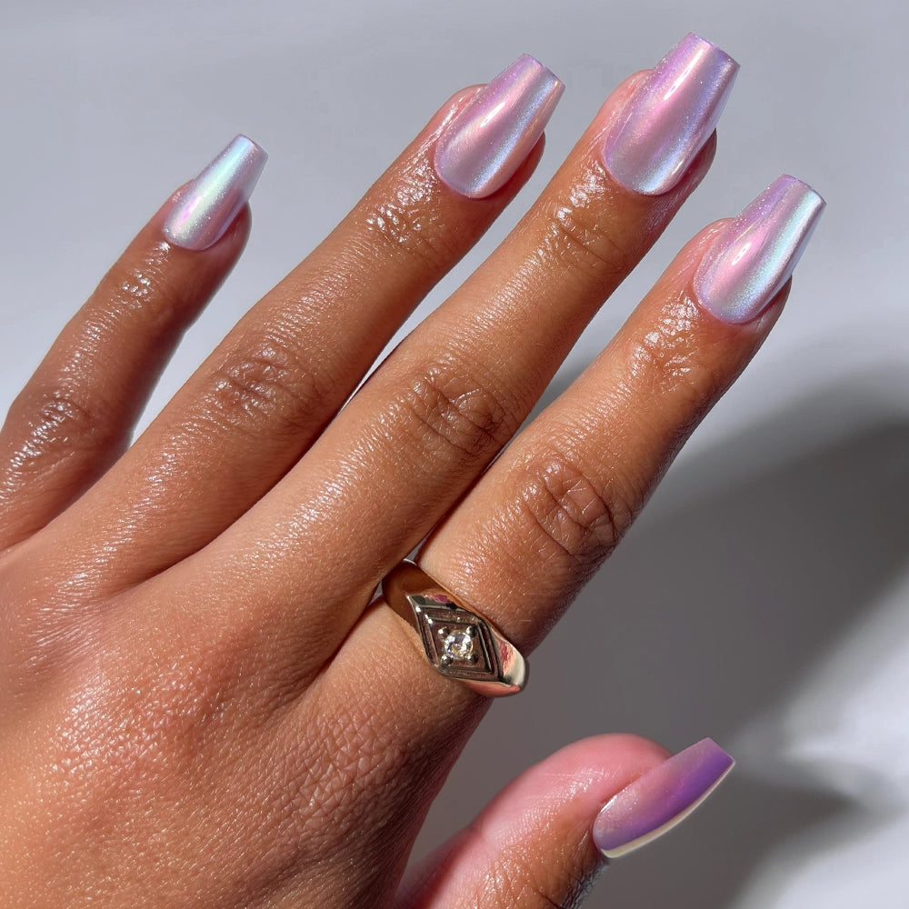 A hand with long, iridescent pink and purple coffin-shaped nails—glamnetic's Oyster Opal set—is shown against a white background. The person wears a gold geometric ring on their middle finger.
