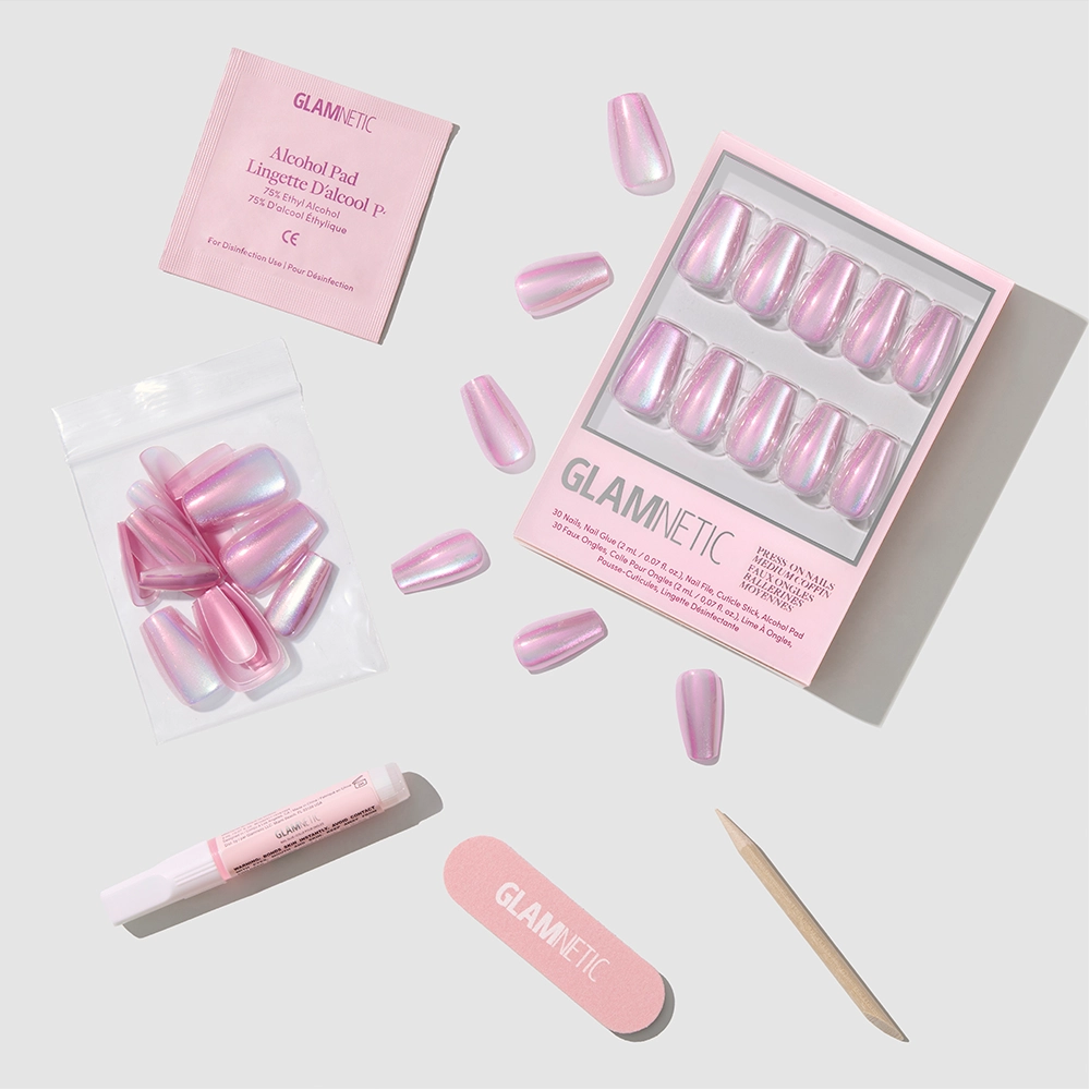 A set of pink iridescent, medium coffin nails from glamnetic's Oyster Opal collection is arranged with an alcohol pad, nail glue, nail file, cuticle stick, and extra nails on a light gray background. The glamnetic logo is on the packaging.