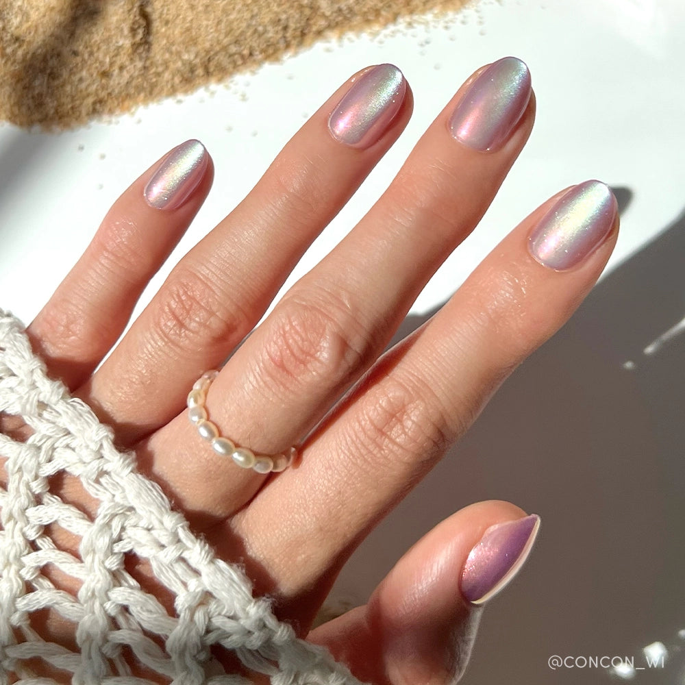 A hand with iridescent pastel rainbow nails from glamnetics Oyster set shines in sunlight. The wearer sports a pearl beaded ring and cream crochet sweater, while the softly lit background highlights the velvet finish.