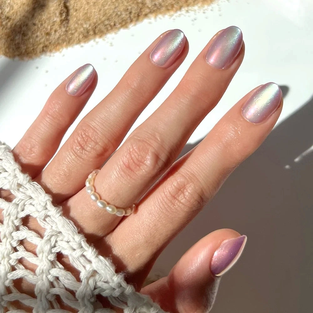 A hand adorned with glamnetic’s Oyster Press-On Nail Set, featuring iridescent pastel pink nails in a short oval shape and paired with a pearl ring, rests on a white surface. The wearer is dressed in a cream crocheted sleeve, bathed in sunlight. These vegan, cruelty-free nails by glamnetic lend an elegant and ethical touch to the delicate look.