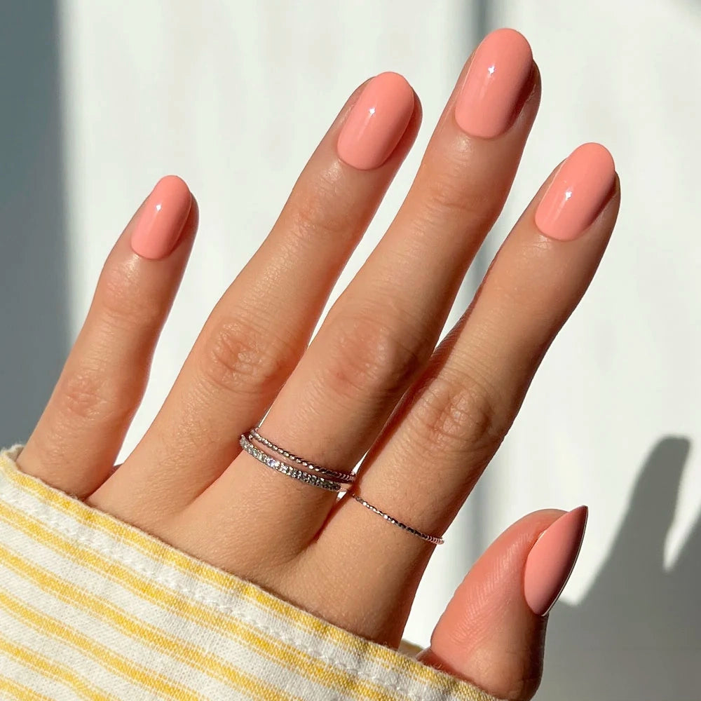 Hand with oval-shaped Glamnetic Peach Coral press-on nails in a glossy finish, wearing two thin metal rings.