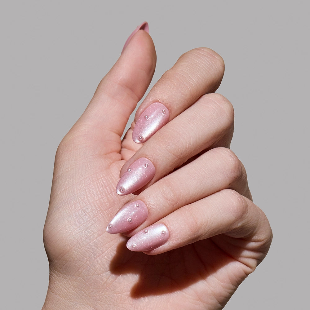A hand with blush-pink almond nails painted in glamnetic’s Pearl Me Softly, a shimmery light pink polish with 3D pearl embellishments, shown against a plain gray background. - Product image
