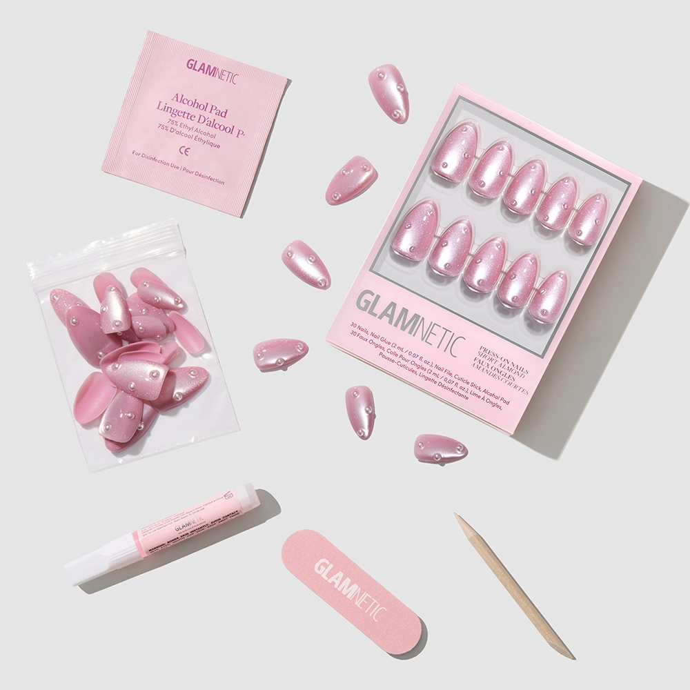 The Pearl Me Softly kit by glamnetic includes blush-pink almond nails with rhinestones and 3D pearls, plus alcohol pad, nail glue, file, cuticle stick, and extra nails—all neatly arranged on a light surface. - Product image