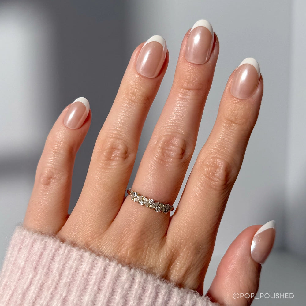 A hand with almond-shaped French tip nails wears glamnetic’s Pearlfect silver ring, featuring five clear gemstones. The person is dressed in a light pink ribbed sweater, with a softly blurred background.