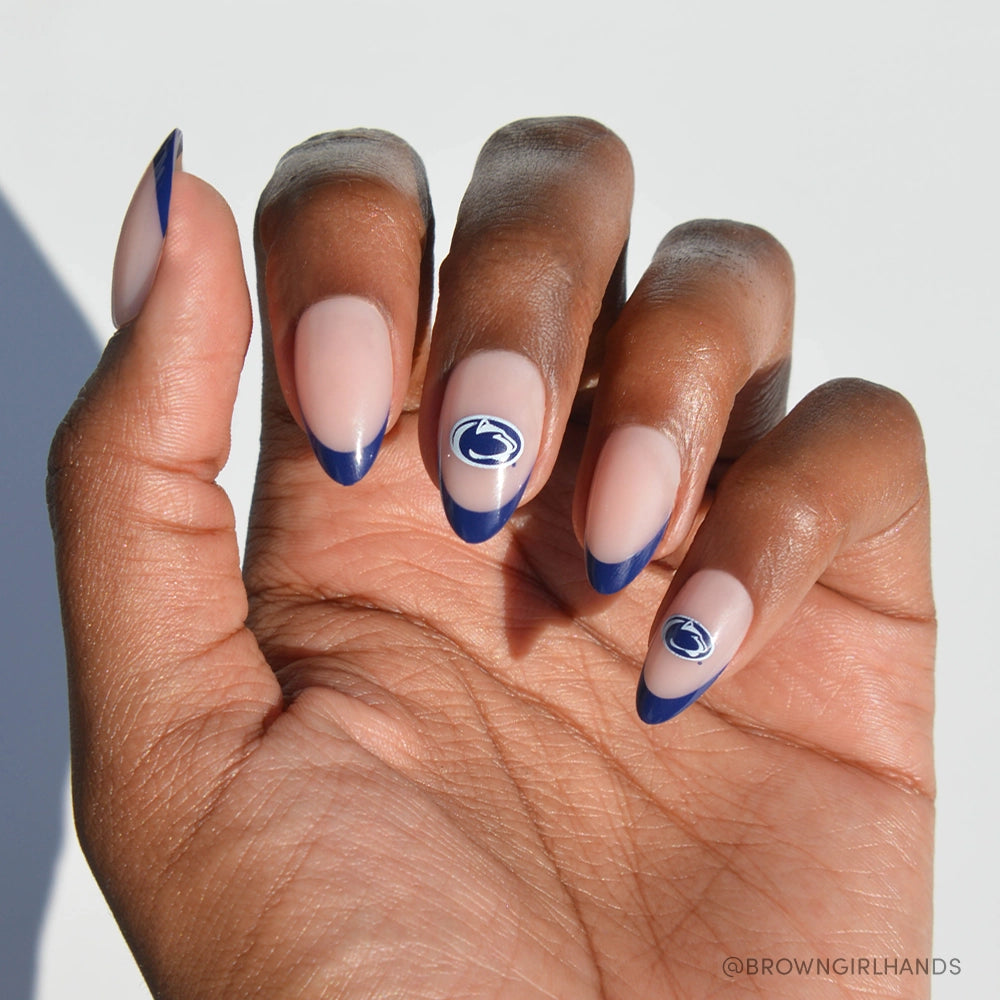 A hand shows almond-shaped nails with a nude base and dark blue French tips, plus two accent nails featuring the blue-and-white Penn State Nittany Lions logo. The glamnetic Penn State Nittany Lions Press-On Nail Set is officially licensed. White background.