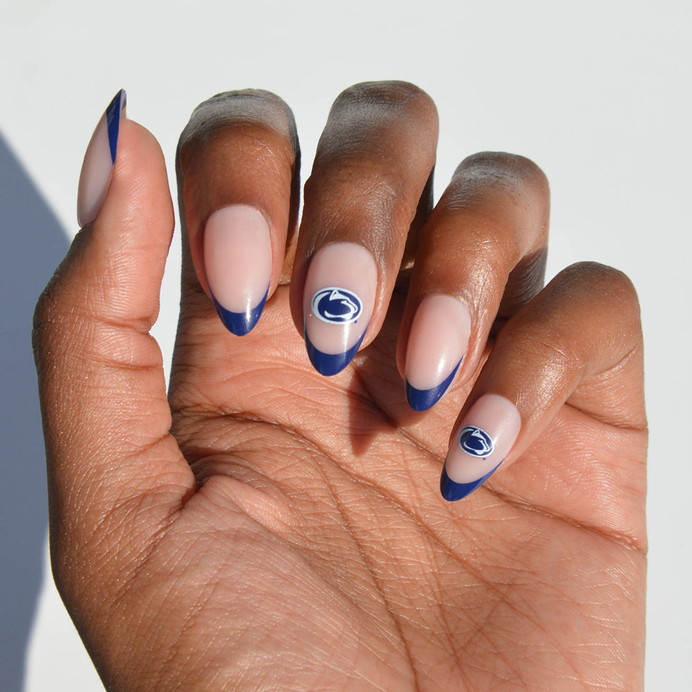 A hand with almond-shaped nails wears glamnetic Collegiate Press-On Nails in a nude shade with dark blue French tips. Two nails are accented with a blue and white paw print logo, set against a plain white background. - Product image