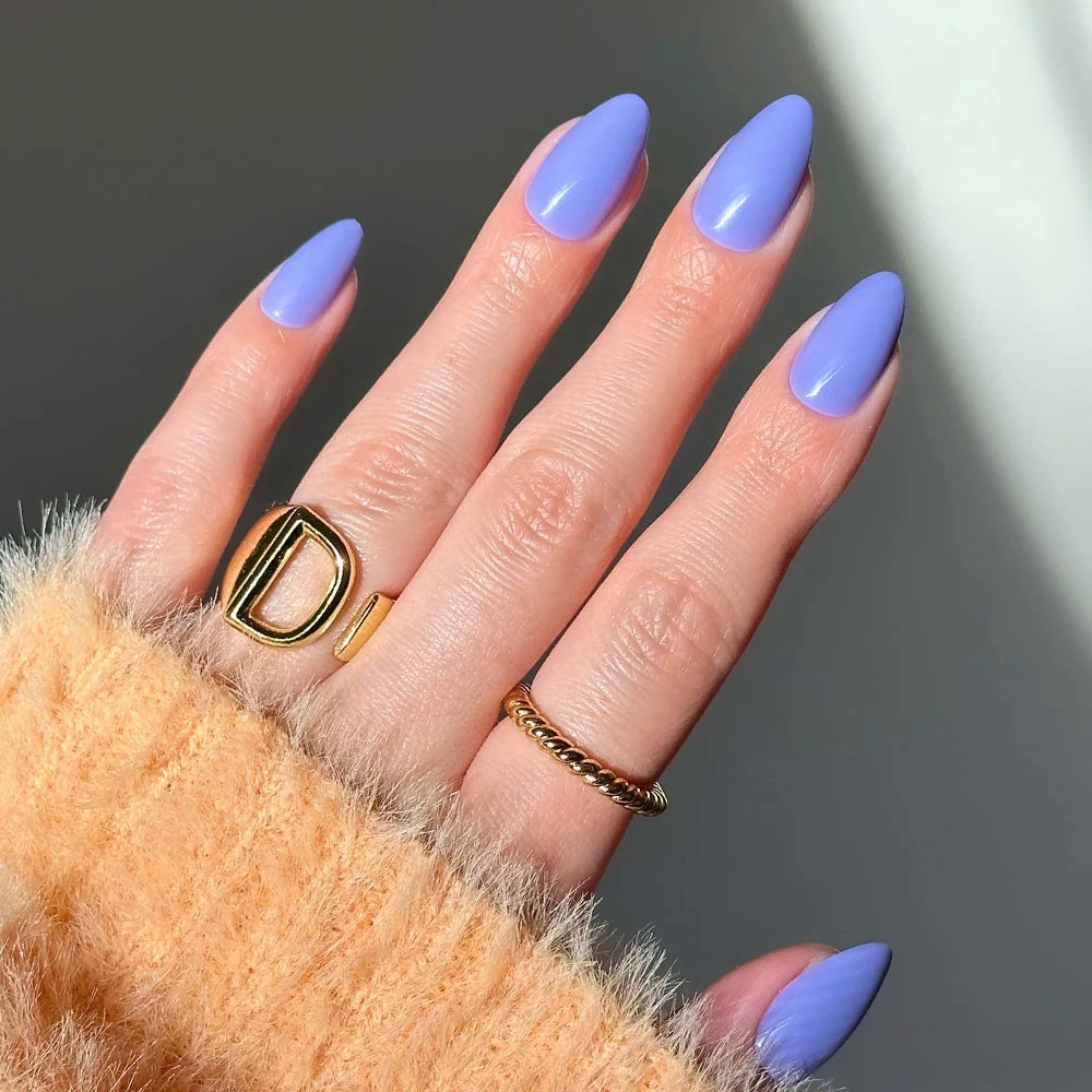 Hand with short almond nails in glossy Periwinkle lilac blue, wearing a gold letter D ring and a thin twisted gold band.