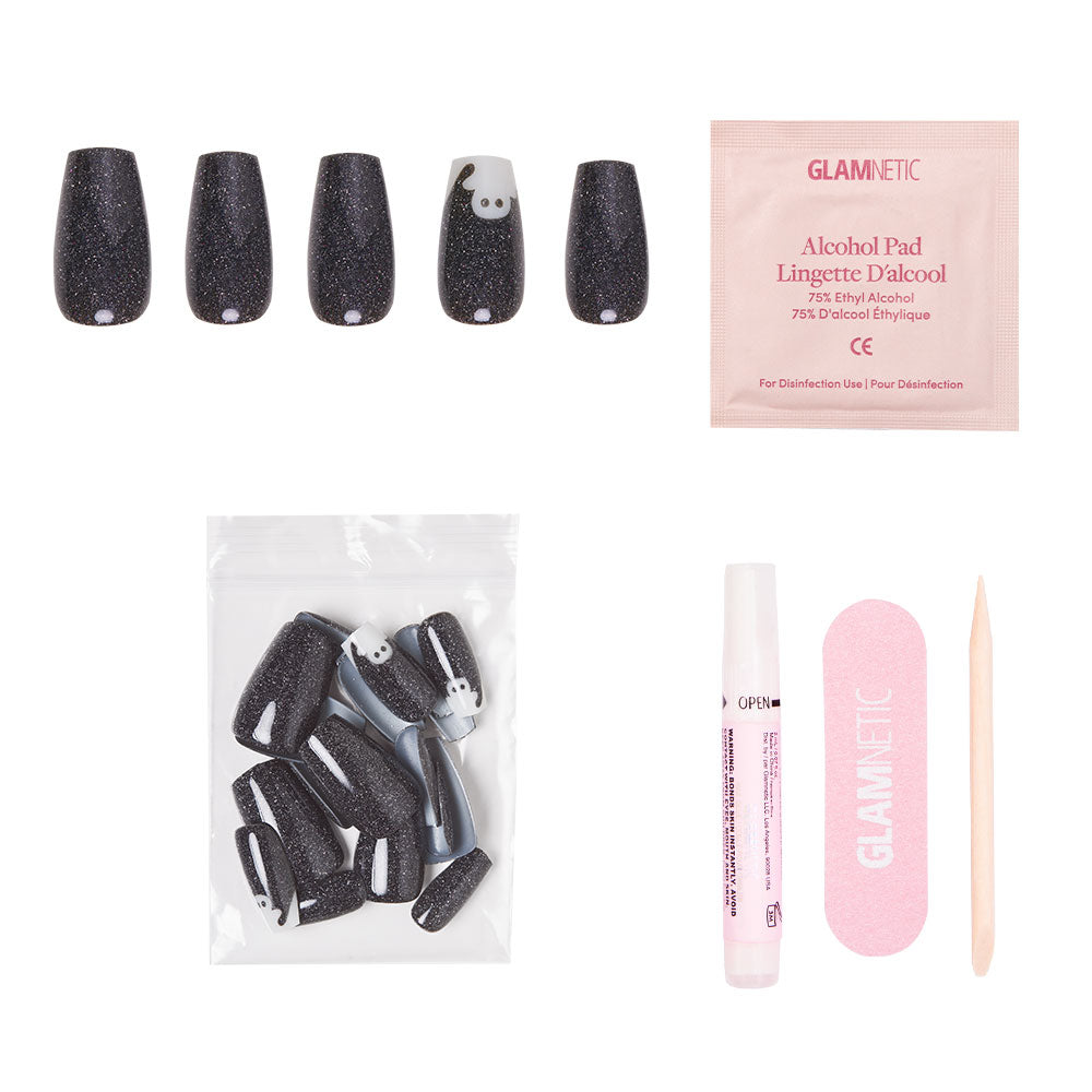 Experience the glamnetic Phantom Glow fake nail kit—a dazzling set of black glitter coffin-shaped press-on nails, highlighted by one unique nail featuring a white cartoon face. The set also includes glow-in-the-dark nails for extra fun, additional nails in a bag, an alcohol pad, nail glue, a pink nail file, and a wooden cuticle stick, all beautifully arranged on a white background. - Product image