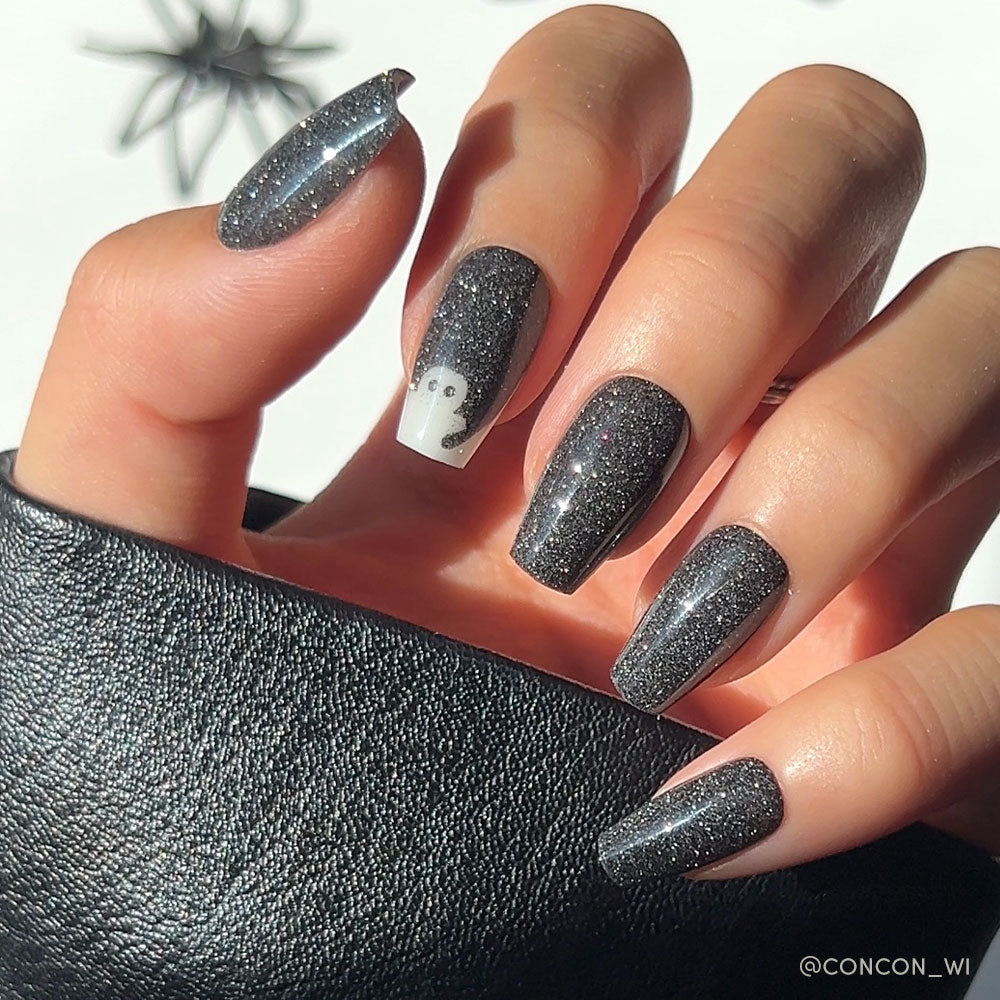 A hand with long, almond-shaped nails painted in sparkly black—just like glamnetic’s Phantom Glow press-on nail set—rests gracefully, the ring finger adorned with a small white ghost design. The hand holds a textured black object, perfectly complementing these glow-in-the-dark nails for an enchanting look.