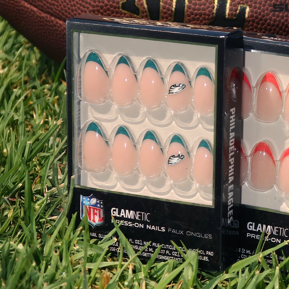 Two boxes of glamnetic Philadelphia Eagles Press-On Nail Sets rest on grass. One set features green and white tips with the Eagles logo, while the other has red and white tips. A football is visible in the background. - Product image