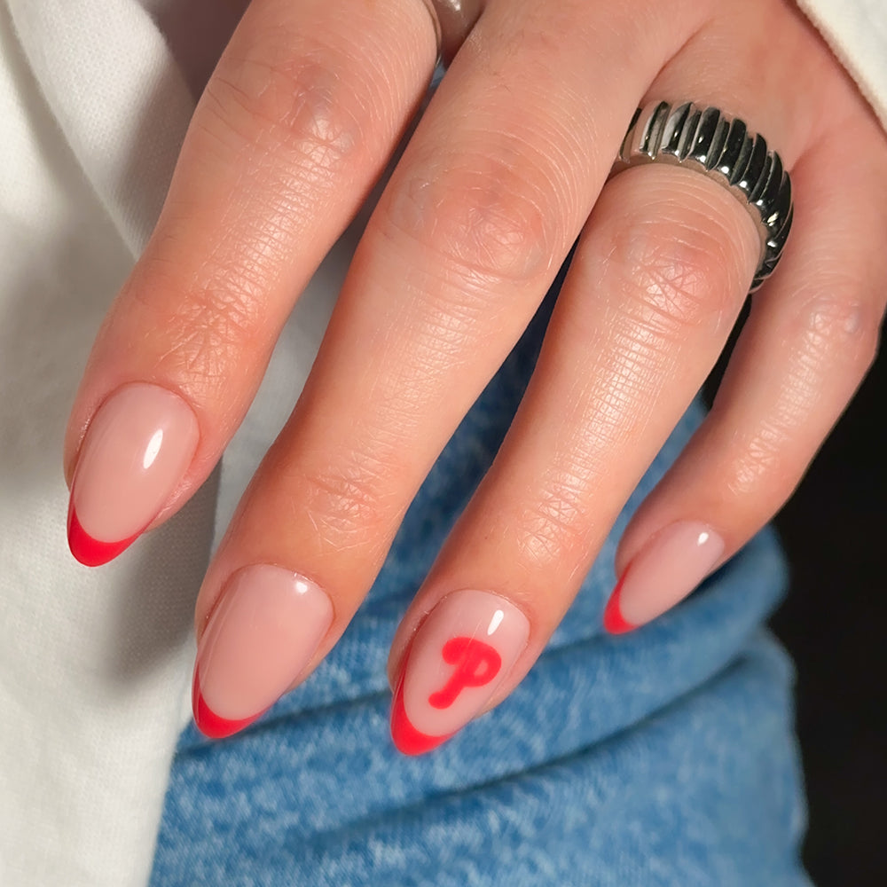 A hand showcases the glamnetic MLB Press-On Nails, styled in an almond shape with a nude base, bold red tips, and a standout red “P” accent on the ring finger. The look is completed with a chunky silver ring and blue jeans. - Product image