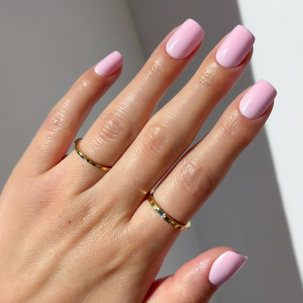 A hand with glossy "Pink Control" by glamnetic on short squoval nails, wearing two thin gold rings on the middle and ring fingers, photographed in natural light against a white background.