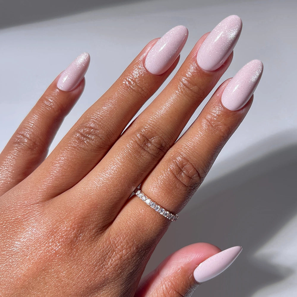 A hand wears glamnetic's Pink Darling salon-quality, light pink, almond-shaped nails and a thin diamond-studded ring on the ring finger, shown in natural light.