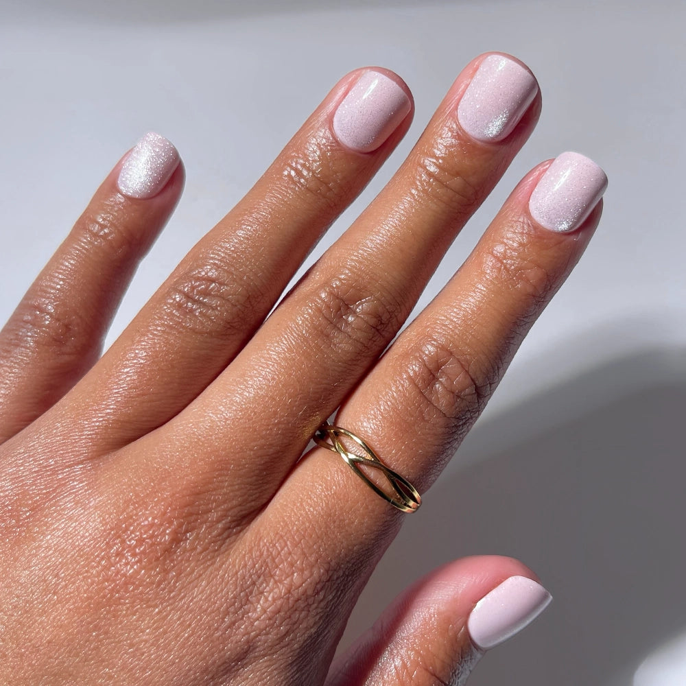 A hand with super short, neatly manicured nails painted with glamnetic's Pink Legacy—a subtle shimmery pink—wears a simple gold ring on the ring finger. The background is white and well-lit.