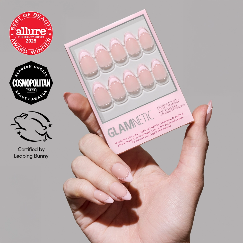 A manicured hand holds a pink box of glamnetic Pink Me Up almond press-on nails with nude, blush pink French tips. Award logos from Allure, Cosmopolitan, and Leaping Bunny are displayed beside the hand.