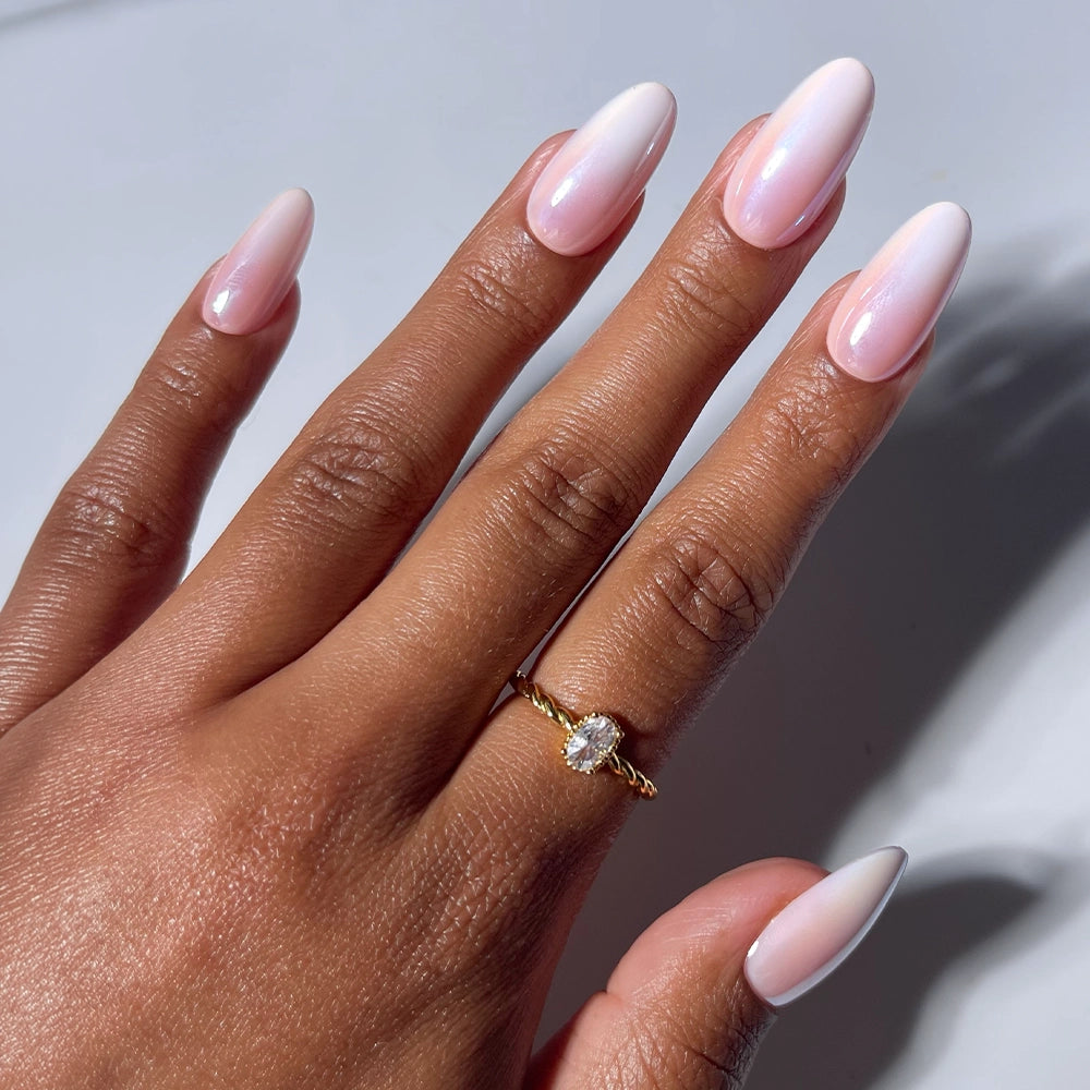 A hand with long, glossy pink ombre French tip nails is shown wearing a gold ring set with a clear oval gemstone. The plain white background highlights the salon-quality Pink Pearl press-on nails by glamnetic.