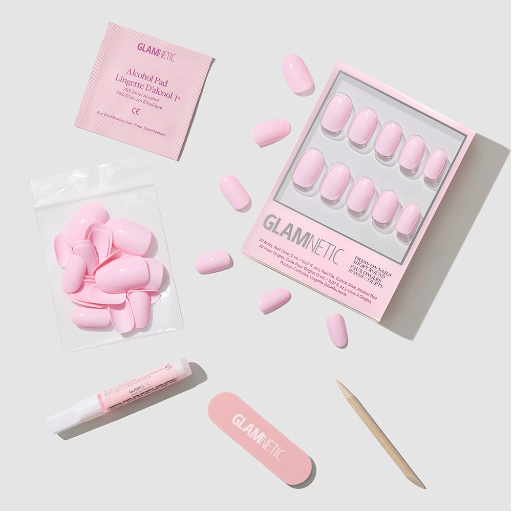 The Pink Permission kit by glamnetic features glossy, short round pink press-on nails with nail glue, alcohol pad, nail file, cuticle stick, and extra nails—all neatly arranged on a light background.