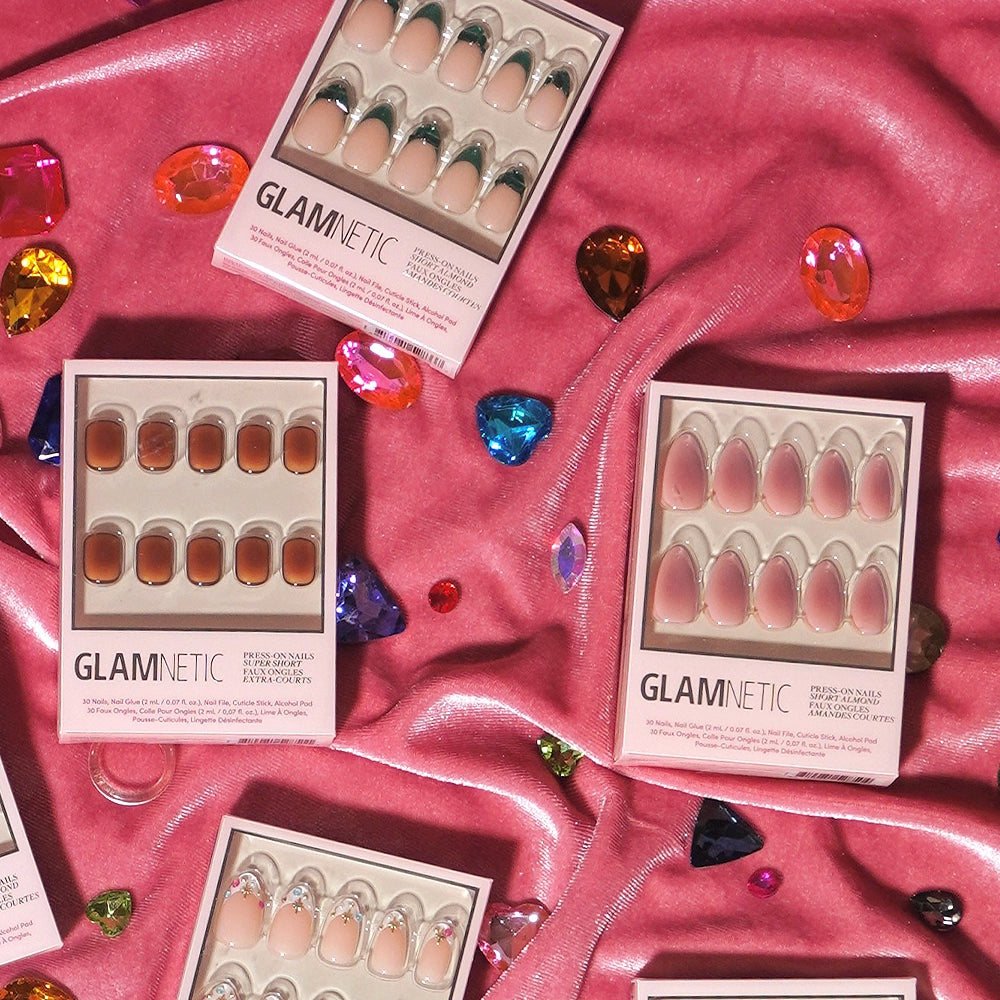 Five boxes of glamnetic press-on nails are arranged on pink fabric with scattered colorful gem decorations. Each box showcases different styles, including chic short almond shapes in nude and brown tones, as well as the elegant Pink Petalite design. - Product image