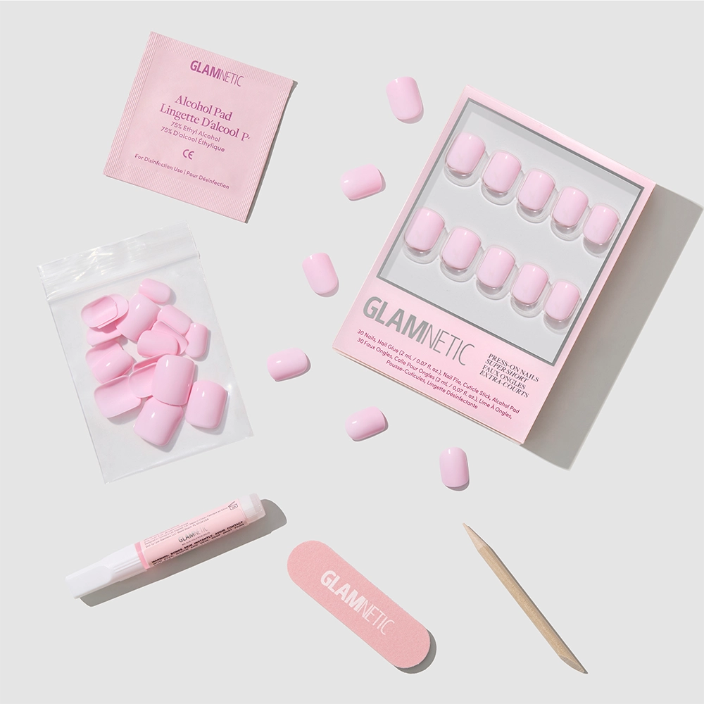 The glamnetic Pink Pop set features glossy blush pink press-on nails, nail glue, a wooden cuticle stick, a pink nail file, extra super short nails in a bag, and an alcohol pad—all arranged on a white background.