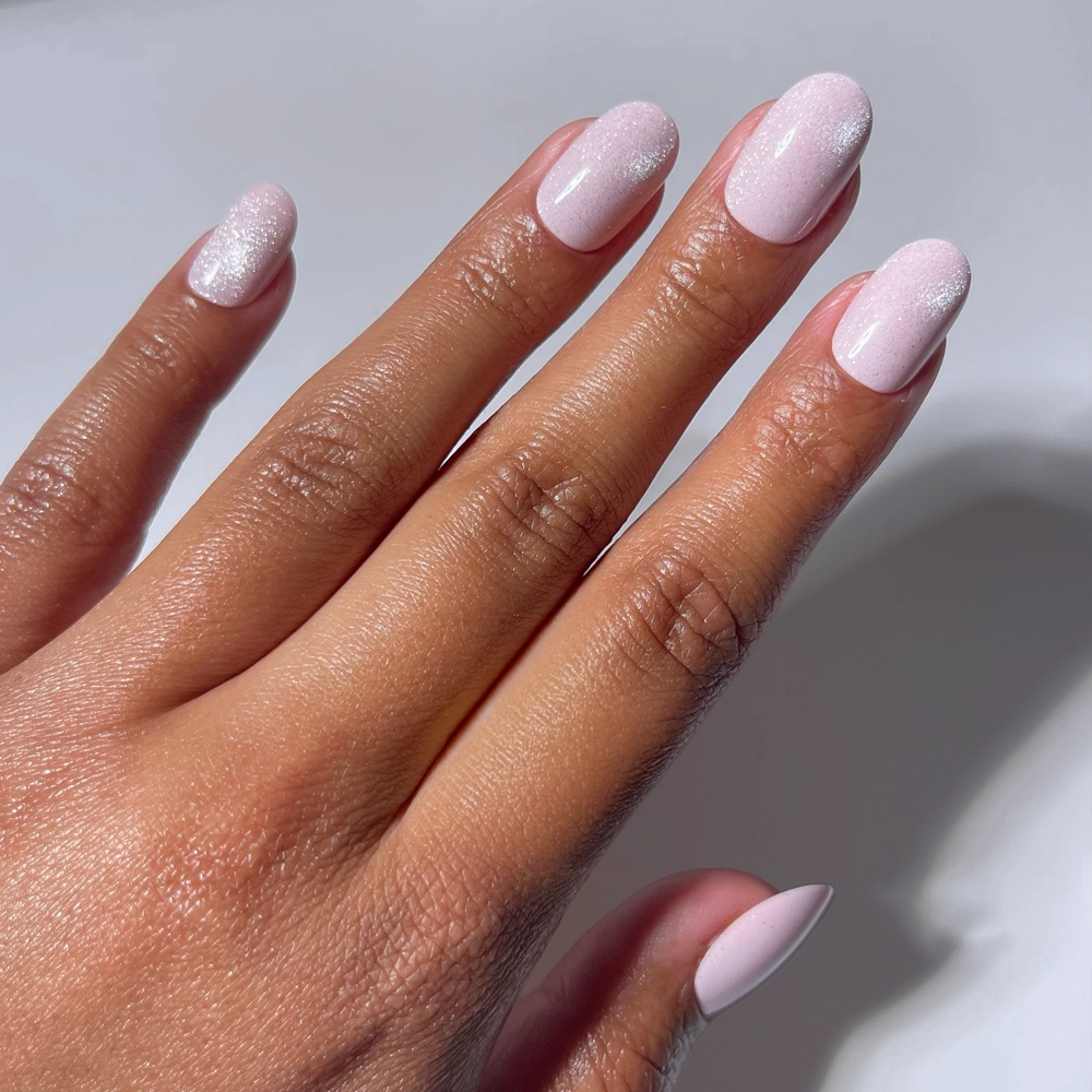 A hand with neatly manicured short round nails showcases glamnetic's Pink Whisper press-ons in light pink, some with a subtle shimmer. The nails stand out against a clean, white background.