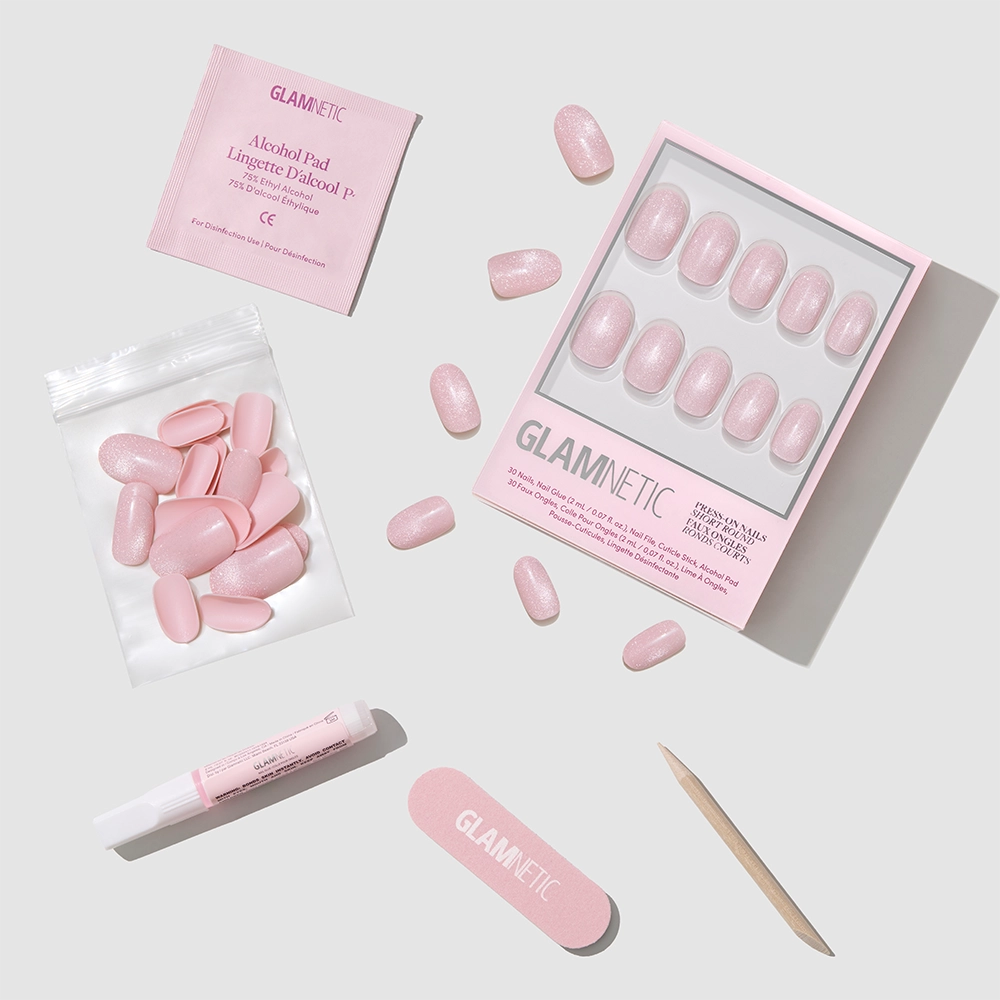 The Pink Whisper set by glamnetic features salon-quality pink press-on nails, glue, alcohol pad, cuticle stick, nail file, and extra short round nails, all labeled “glamnetic” and displayed neatly on a light gray surface.