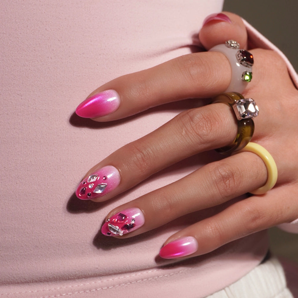 A hand adorned with glamnetic’s Pink Tourmaline press-on nails—long, pink ombre nails embellished with rhinestones and gemstones. The person pairs multiple chunky rings and a light pink long-sleeve top to perfectly complement the glamorous set. - Product image