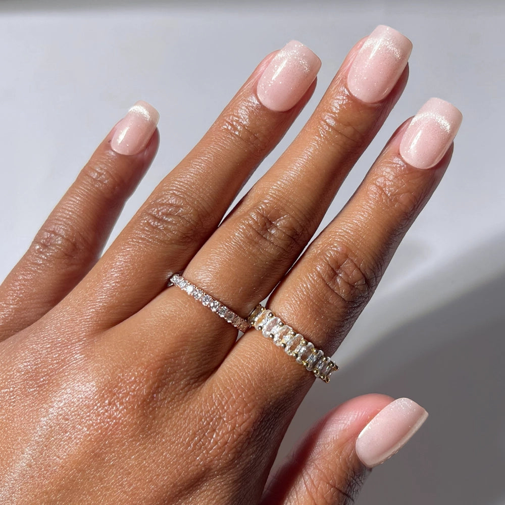 A hand with short squoval nails painted in “Pixie Dusted” by glamnetic—a light pink, glossy shade—wears two gold rings adorned with small diamonds on the ring and middle fingers against a light background. - Product image