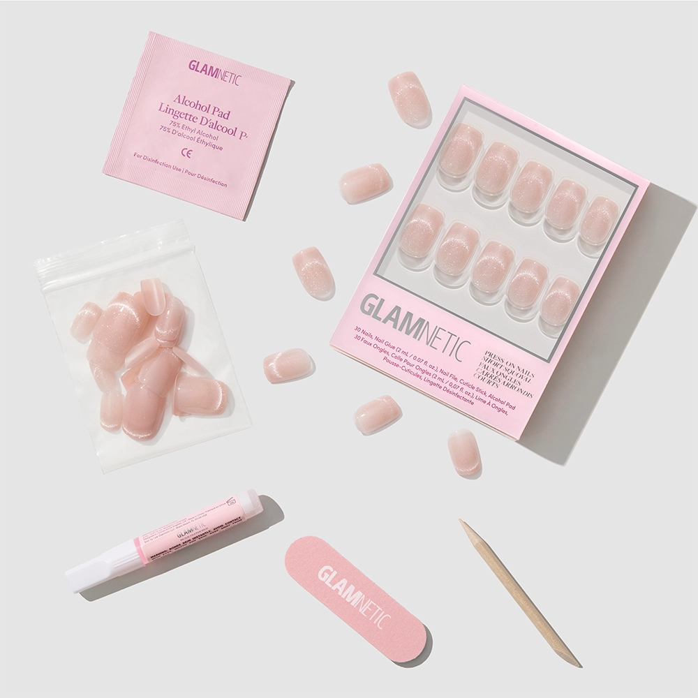 The Pixie Dusted press-on nails by glamnetic—a set of pink, short squoval nails featuring a subtle cat-eye finish—are displayed neatly on a light surface alongside nail glue, an alcohol pad, a nail file, a cuticle stick, and extra nails. - Product image