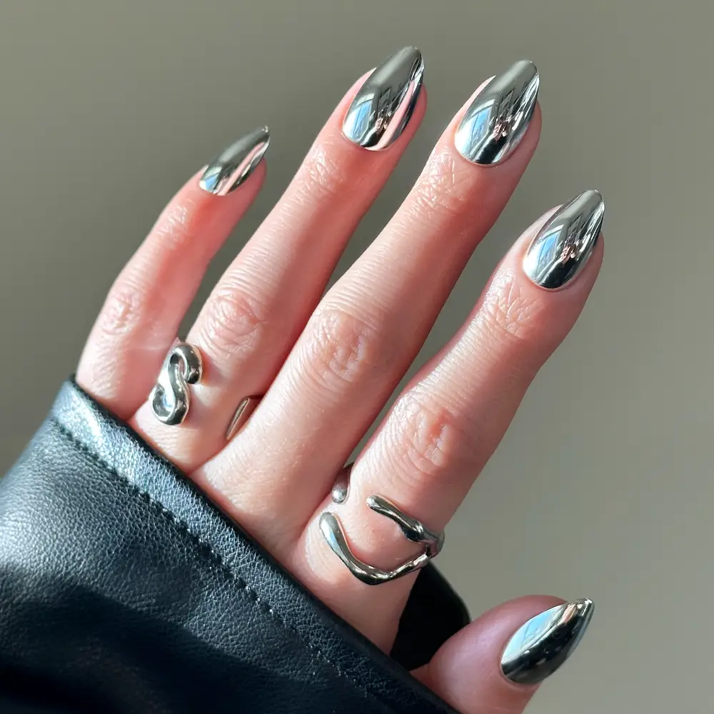 A hand with glamnetic Platinum chrome almond-shaped nails, adorned with two chunky silver rings and a black leather sleeve partially visible.