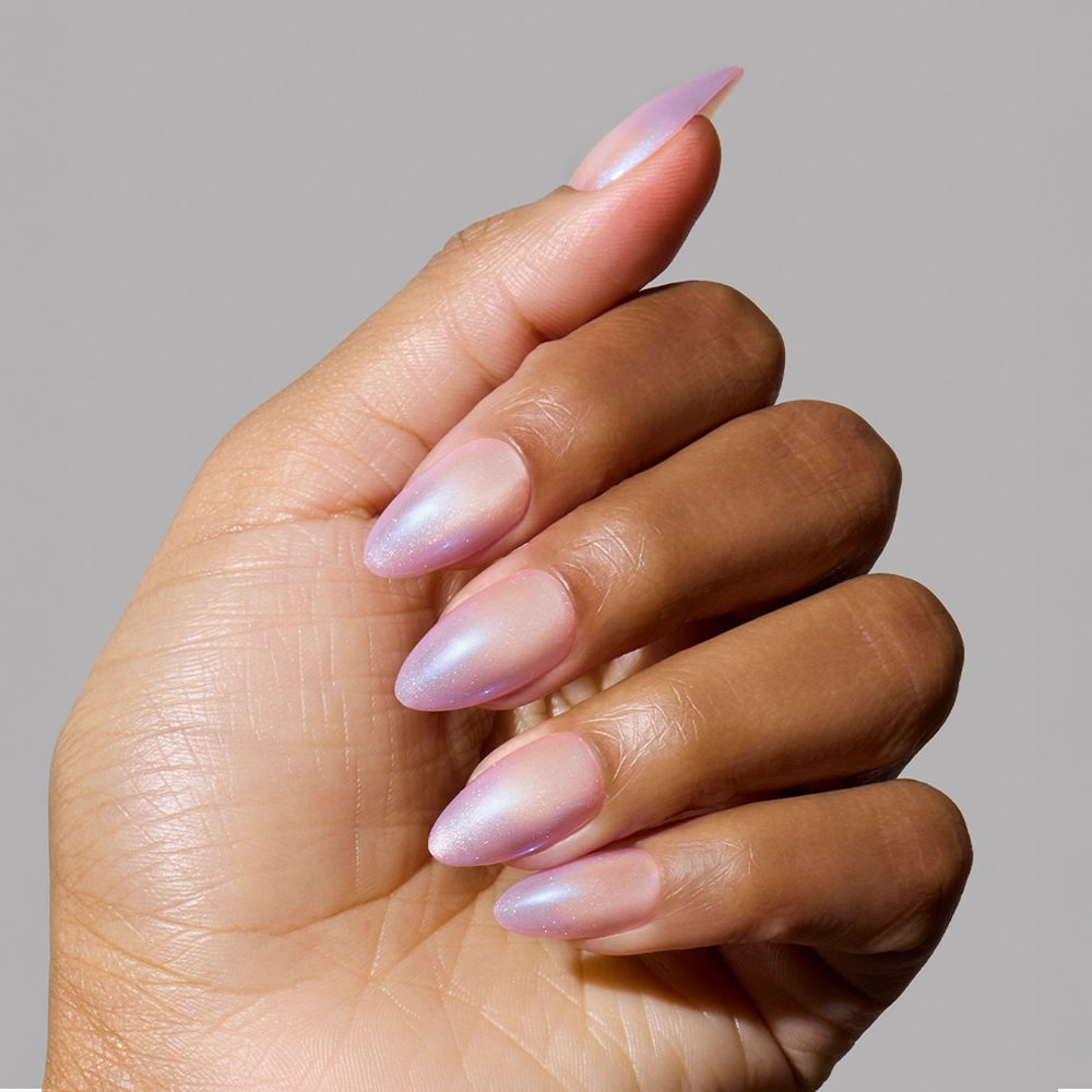 A medium-skinned hand displays glamnetics Polar Day minimalist nail set with short almond nails in glossy, iridescent light pink polish against a plain gray background. - Product image