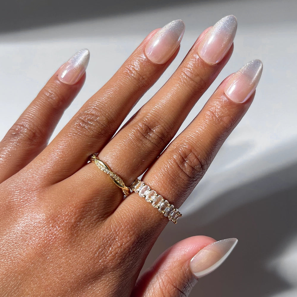 A hand adorned with long, almond-shaped, gradient nude press-on nails from the glamnetic Polar Night set is styled with two rings: a twisted gold band and another gold ring featuring rectangular clear stones.