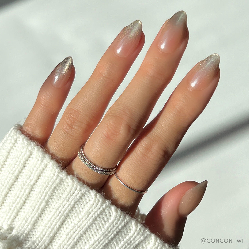 A hand with long almond nails in a nude-to-silver ombre and glossy velvet finish models glamnetic’s Polar Night set, paired with two minimalist rings and a white knit sweater against a soft light background. - Product image